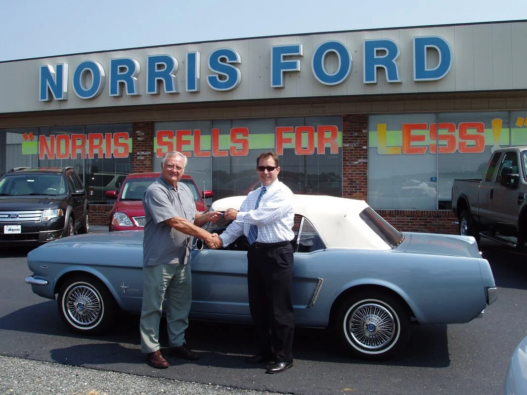 Two men shaking hands in front of a vintage convertible car outside a dealership with a sign that reads "NORRIS FORD", and a banner stating "NORRIS SELLS FOR LESS!"