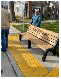 A park bench with a plaque that reads 'Donated by the class of 1974, Warm Sand' on a sidewalk with yellow tactile paving. Two men are standing nearby, one wearing a blue jacket and hat.