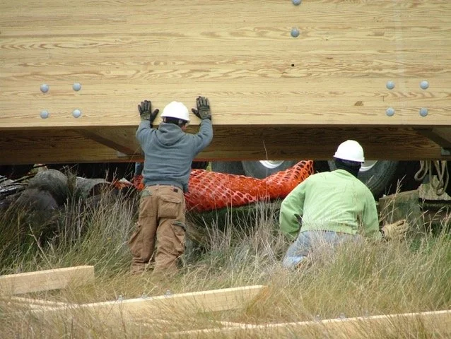 Two construction workers wearing helmets and gloves installing a large wooden panel over a red dinosaur statue and some tires at a construction site with grass and wooden planks.