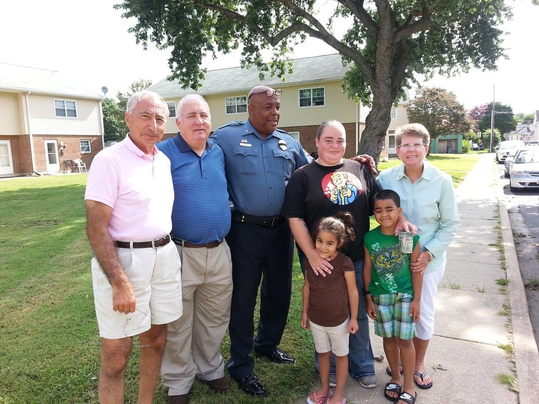 A group of seven diverse people, including children and adults, standing on a sidewalk in front of a house and tree. They appear to be smiling and enjoying a day outside.