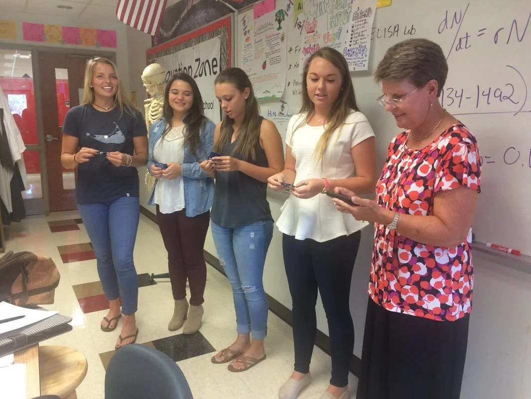 A teacher and four teenage students are standing in a classroom, all looking at their phones.