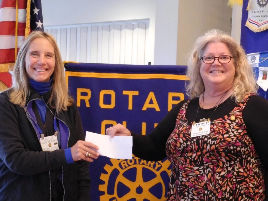 Two women smiling and exchanging an envelope in front of a blue and gold Rotary Club banner. One woman has blonde hair, is wearing a black jacket and name tag that reads 'Julie Andrews,' and the other woman has curly blonde hair, glasses, and a color