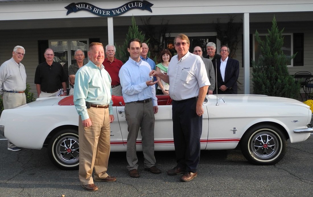 A group of men standing in front of a white vintage convertible car with red accents, at a yacht club event. Two men in the front are exchanging an object, possibly a trophy or award. The background features a building with a sign that reads "Miles R