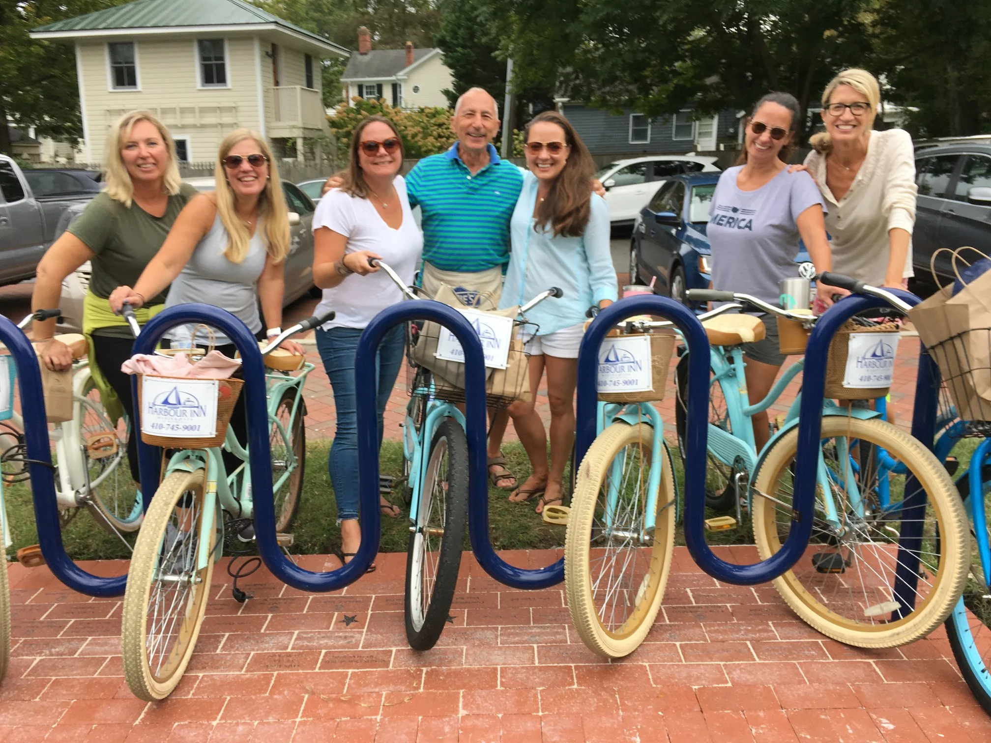 Group of eight smiling women and one man standing behind bicycles with front baskets, in an outdoor area with parked cars and houses in the background.
