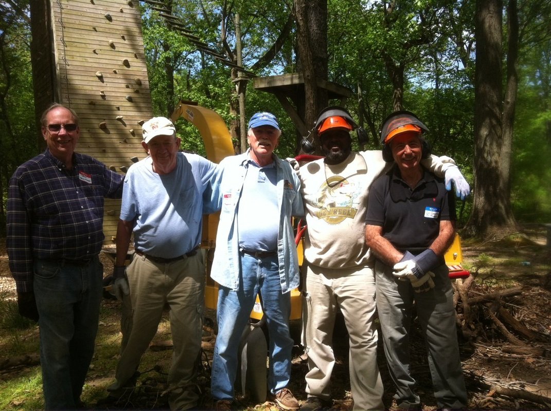 Five men standing outdoors in a wooded area. They are wearing work gloves and safety helmets, with one man in the background standing next to a large piece of wood and a small yellow construction machine.