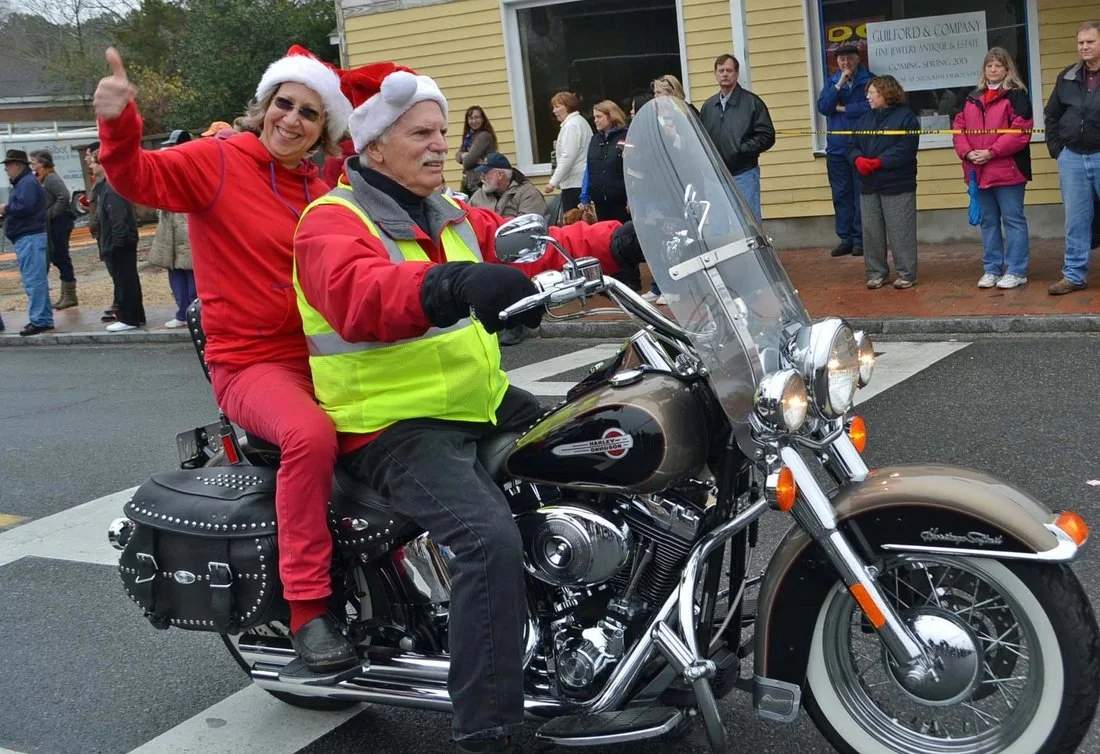 A man and a woman wearing Santa hats riding a Harley-Davidson motorcycle during a holiday event. The woman is giving a thumbs-up and smiling, while the man is focused on riding. Spectators are watching from the sidewalk.