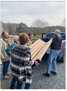 Group of people unloading a wooden board from a blue pickup truck outdoors on a cloudy day.