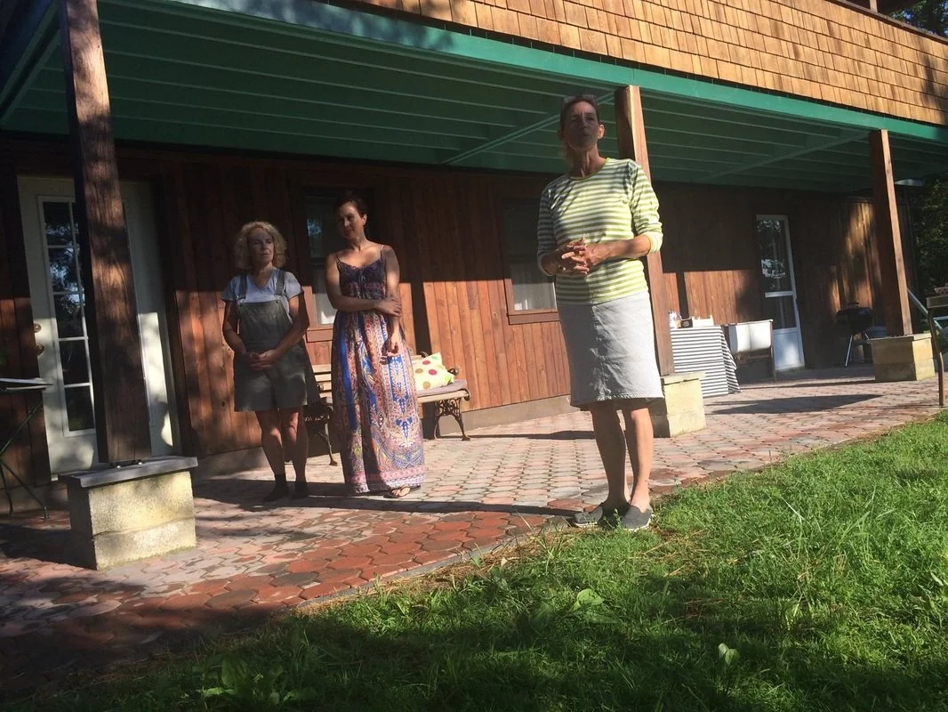 Three women standing on a brick patio outside a wooden house in sunlight. One woman in a striped shirt and white skirt is in the foreground, while the other two women stand behind her, one in overalls and the other in a colorful dress.