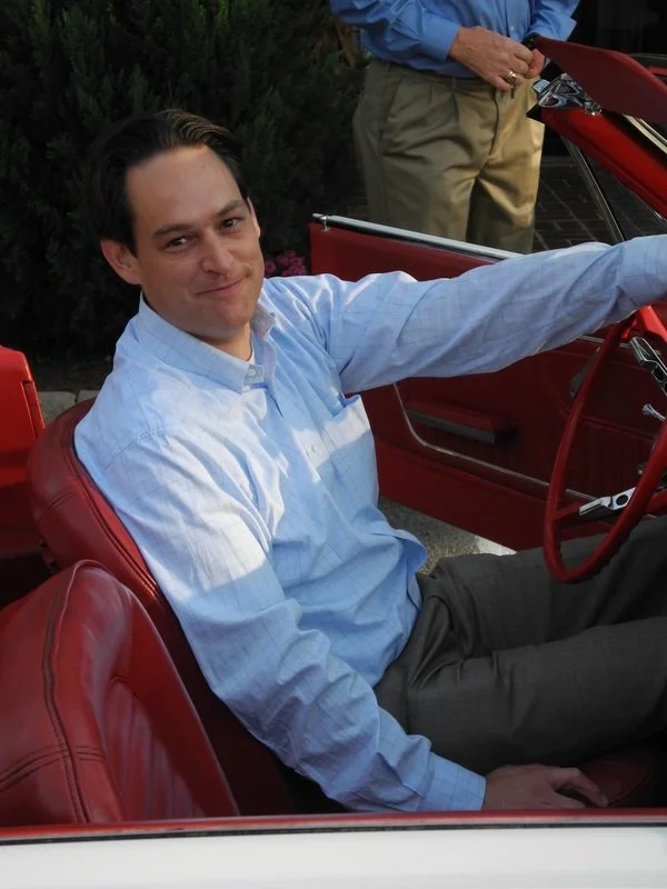 A man sitting in the driver's seat of a vintage red convertible car, smiling and reaching out with his right arm. Another person is standing next to the car wearing beige pants and a blue shirt. The background features greenery.