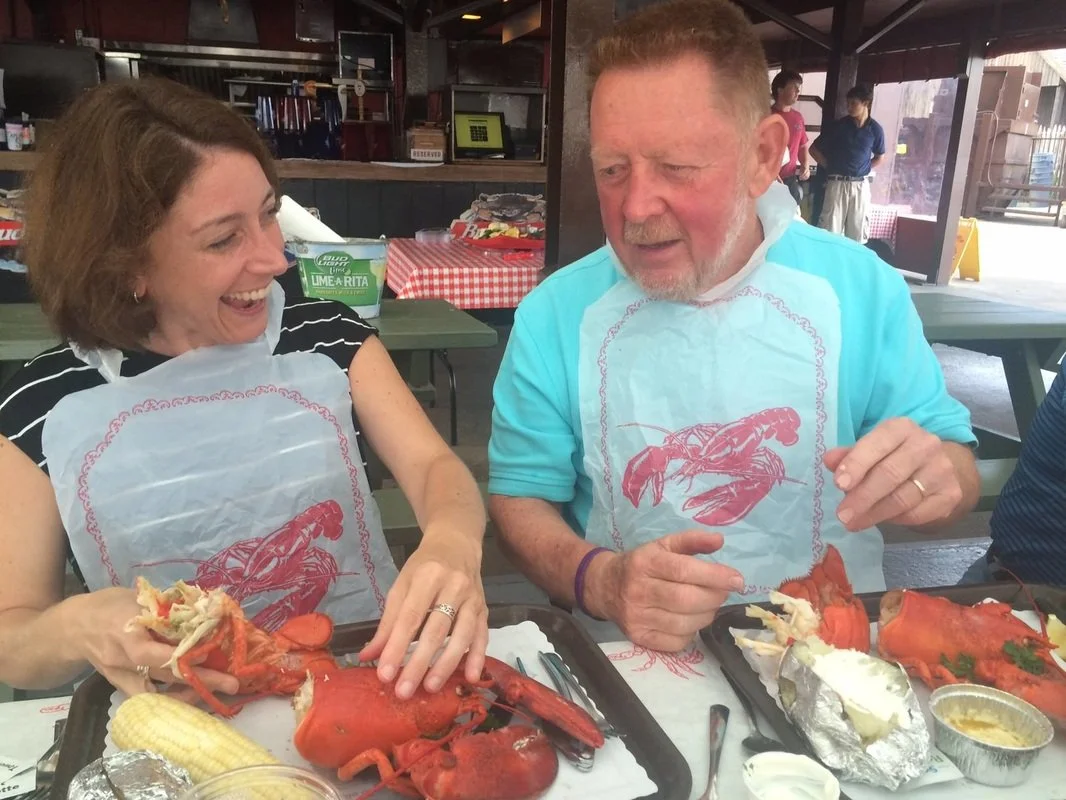 Two people enjoying a seafood meal at a restaurant, wearing bibs with lobster illustrations, with lobsters, corn, and potato on trays in front of them.