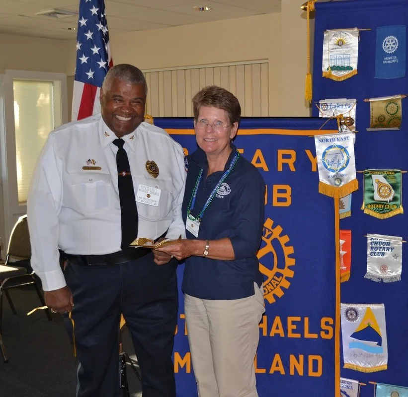 A man in a white police uniform with a black tie smiling and holding a certificate with a woman in a blue jacket and beige pants, both standing in front of a blue Rotary Club banner and various banners, with an American flag in the background.