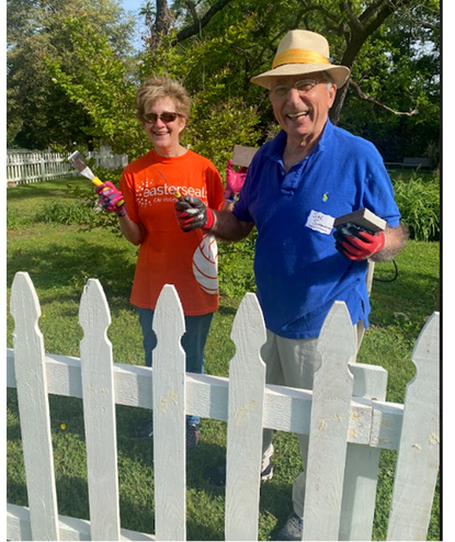 Two smiling people, an older man wearing a straw hat and blue shirt, and a woman in an orange shirt, painting and working on a white fence in a backyard with trees.