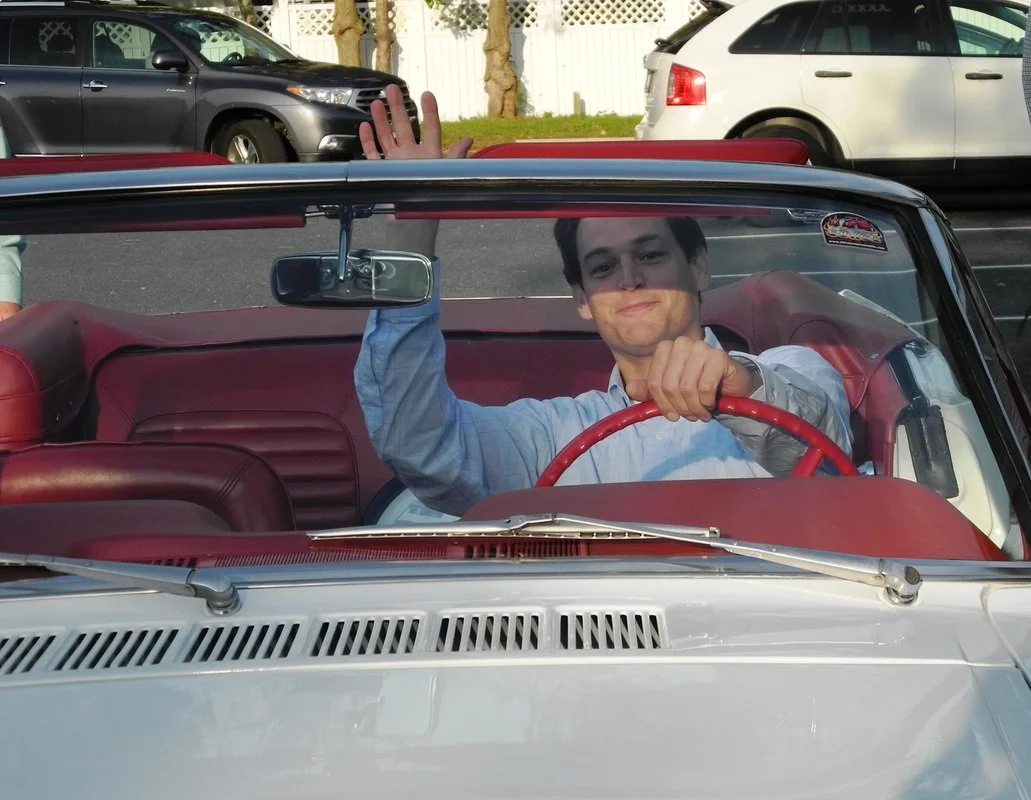 A young man in a light blue shirt sits in the driver's seat of a vintage convertible car with red interior, smiling, and waving at the camera. The car has a clear windshield and is parked outdoors with other vehicles and trees visible in the backgrou