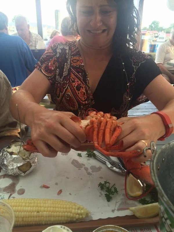 Woman opening a cooked lobster at a restaurant table, with corn on the cob, lemon, and other table items visible.
