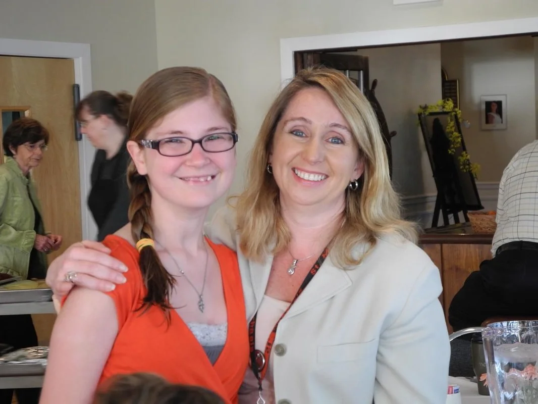 Two women smiling, one with glasses and red hair in a braid, the other with blonde hair, at an indoor social event.