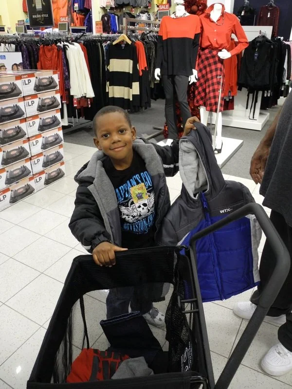 A young boy shopping in a clothing store, holding a backpack near a shopping cart, with clothing displays and mannequins in the background.