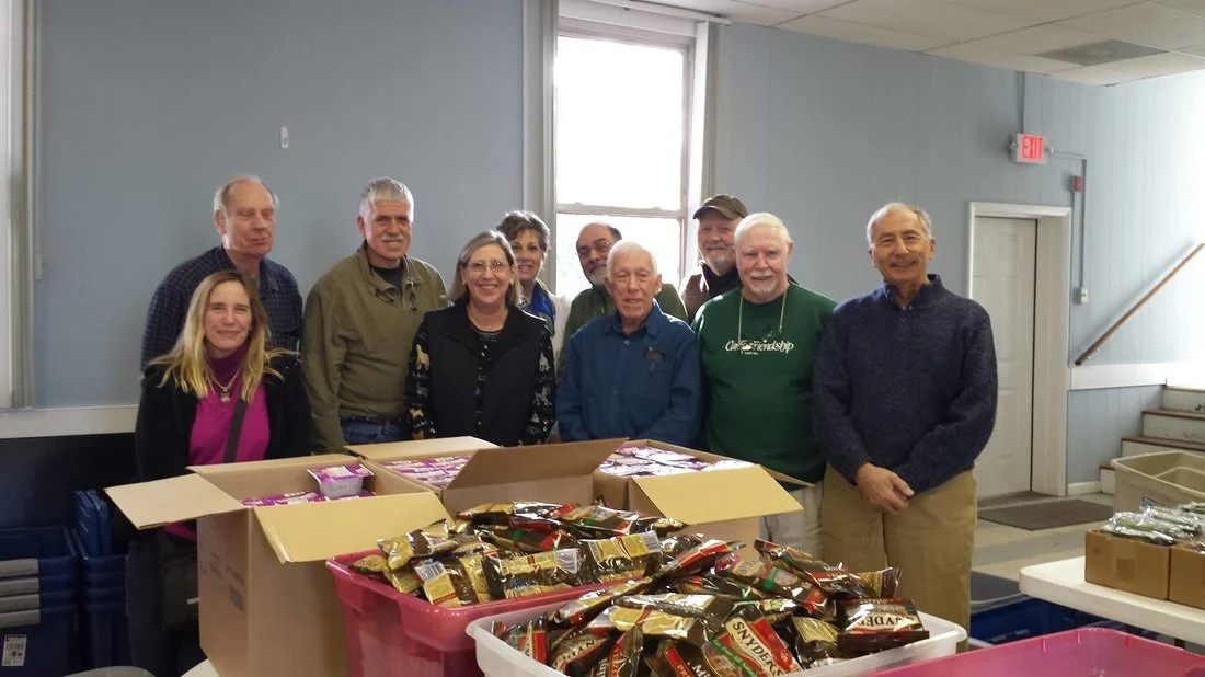 Group of ten people standing behind tables with boxes and packages of food, inside a room with light blue walls and a window.