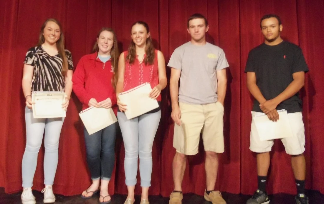 Group of five young people standing in front of a red curtain, holding certificates, at an award ceremony or graduation event.