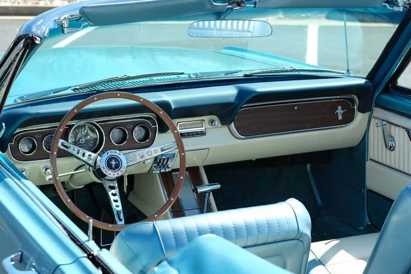 Interior of a vintage blue Ford Mustang car, showing the dashboard with wood and chrome accents, a classic steering wheel with the Ford Mustang logo, and white leather seats.