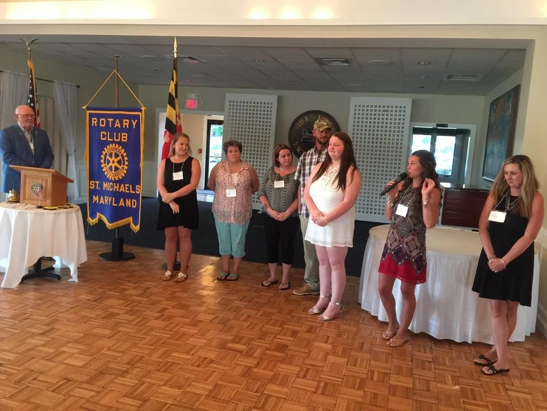 A group of people standing in a line at a Rotary Club event, with a man at a podium and a Rotary banner in the background. The setting appears to be a banquet or meeting room.