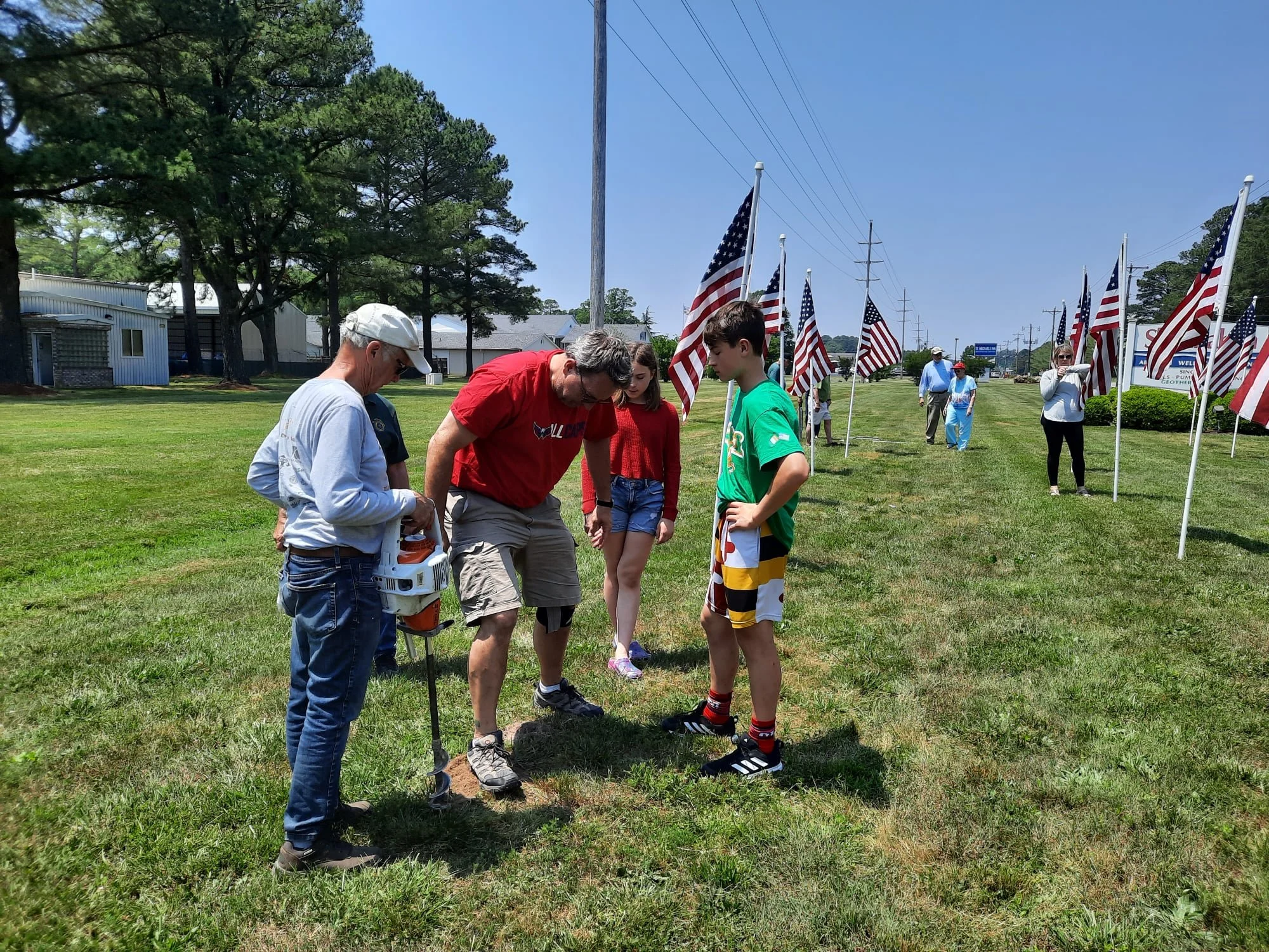 People planting a tree on a grassy lawn with American flags on poles along the sidewalk.