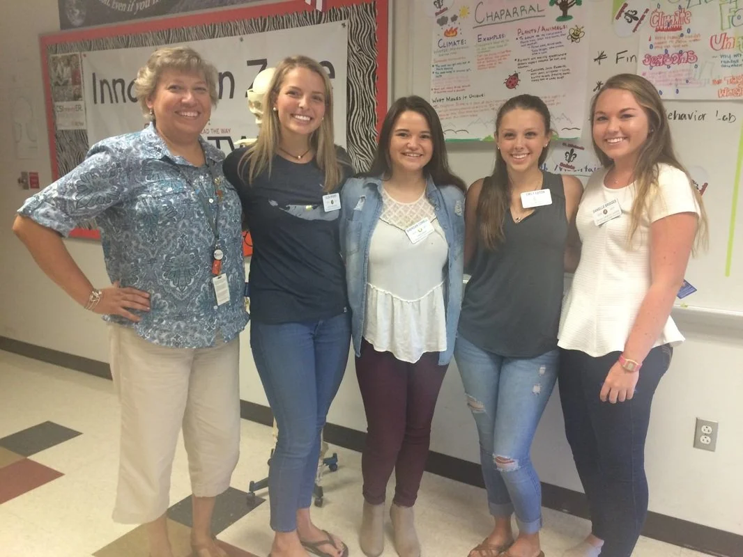 Group of five women standing together in front of a whiteboard with colorful posters and notes, smiling at the camera.
