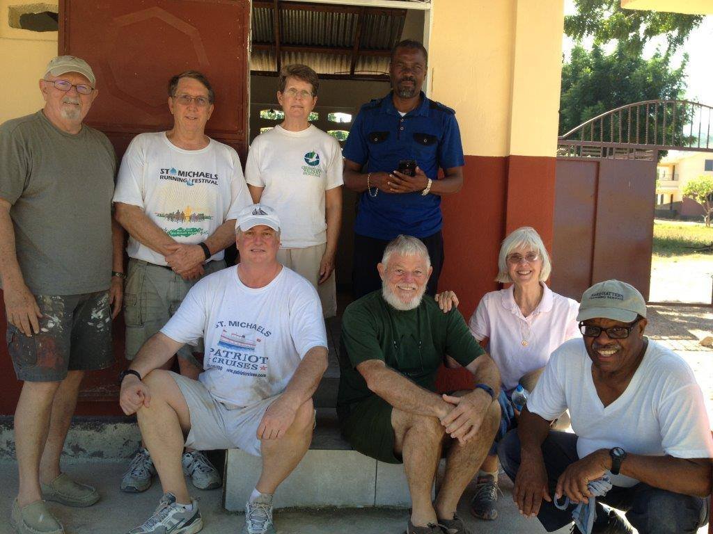 A group of nine people, mostly seniors, pose together outdoors in front of a building with trees in the background. They are smiling and dressed casually, some wearing hats and athletic wear.