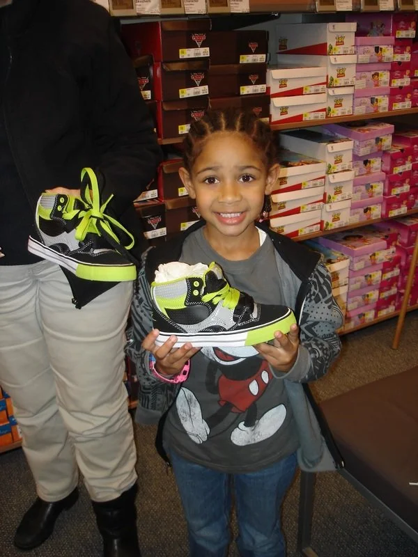 A young girl with braided hair holding a neon green and black Nike sneaker inside a shoe store, with shelves of shoes behind her.