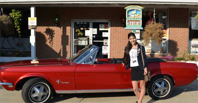 A woman standing next to a red vintage convertible car parked in front of a brick building.
