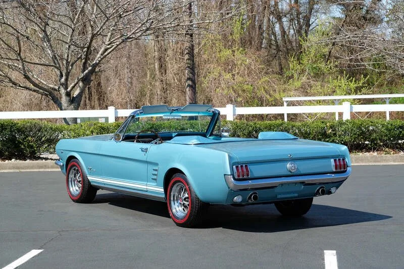 A light blue vintage Ford Mustang convertible parked on an empty parking lot with a white fence and trees in the background.