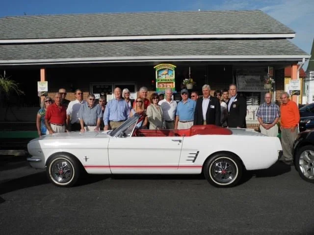 Group of people gathered behind a white convertible car with red interior parked in front of a building during daytime.