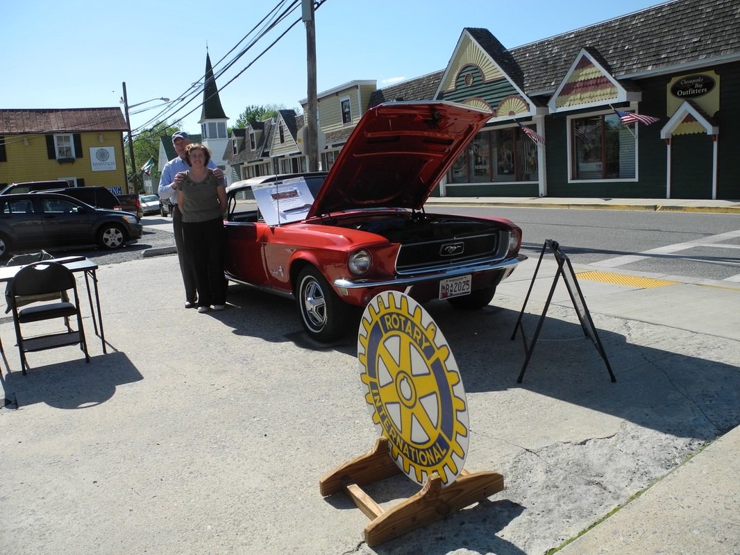A classic red Ford Mustang muscle car with its hood open, displayed outdoors with a Rotary International sign nearby, on a street with storefronts and people observing.