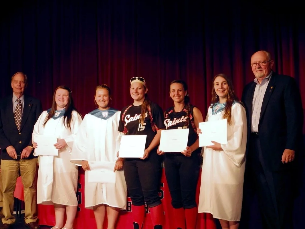 Group of six young women and two older men standing on stage with red curtains. The women are holding certificates and wearing sports uniforms or white robes, indicating an award ceremony or recognition event.