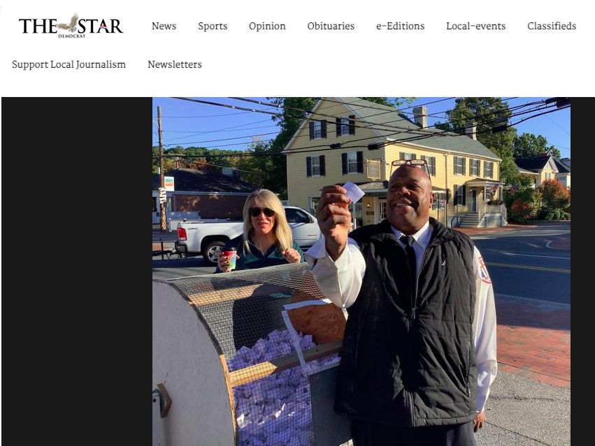 A man in a uniform and a black vest smiling and holding a small piece of paper or ticket, standing outside on the sidewalk. Behind him, a woman with blonde hair, sunglasses, and a dark jacket stands near a street corner, with a white truck and reside