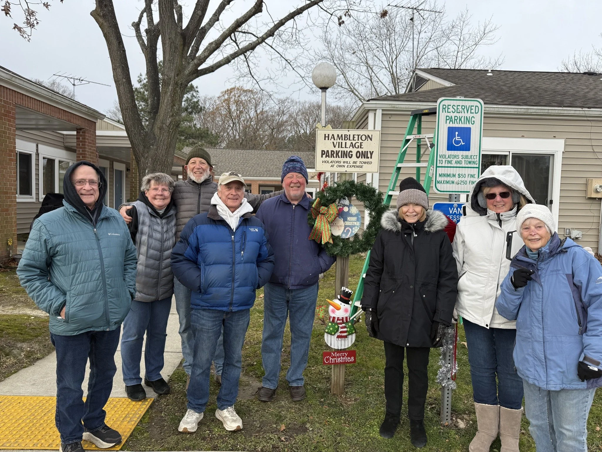 Group of nine people standing outdoors near parking signs decorated with Christmas wreath, snowman, and holiday decorations, wearing winter jackets and hats.