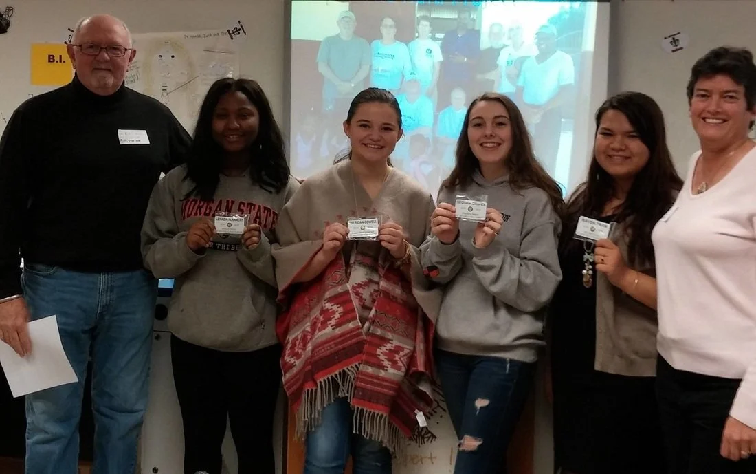 Group of six people, including five young women and one older man and woman, standing in a classroom, holding certificates, with a projection of a group photo on the wall behind them.
