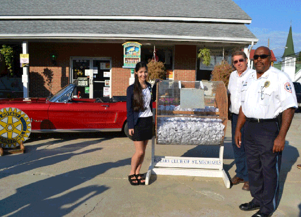 A girl and two men standing outdoors near a Rotary Club of St. Michaels booth with ice, a vintage red convertible car, and a building in the background.
