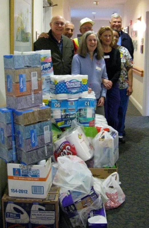 Group of six people standing behind a large collection of donated household supplies, including paper towels, tissues, and cleaning products, in a hallway.