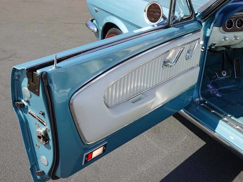 Close-up of a vintage light blue car door interior showing silver trim, a door handle, window crank, and speaker, with part of the car's dashboard and another classic car in the background.
