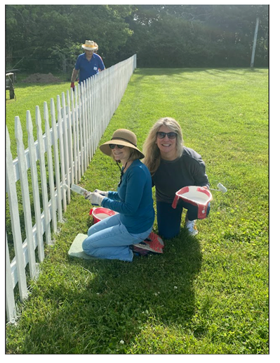 Two women sitting on the grass painting a white picket fence, with one woman wearing a hat and gloves, and two people gardening in the background.