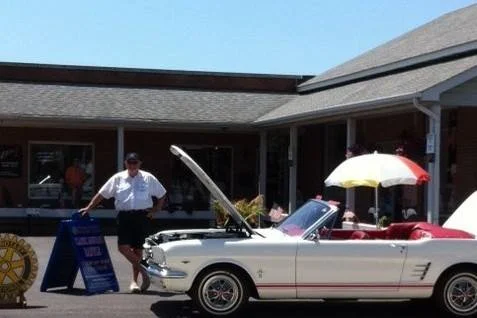 A man standing next to a white vintage convertible car with a raised hood, parked in front of a building with an awning, under a sunny sky, with a yellow and white umbrella and a signboard nearby.