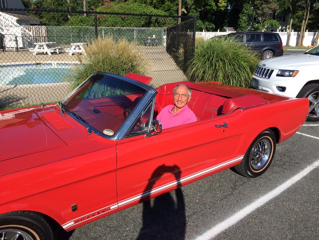 A smiling older man sitting in a red vintage Ford Mustang convertible parked on a street, with a tennis court and parked cars in the background.