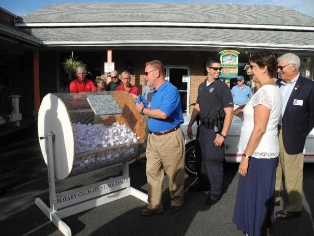 People gathered around a large raffle drum labeled 'Rotary Club of St. Michael's,' participating in a drawing event outside a building.