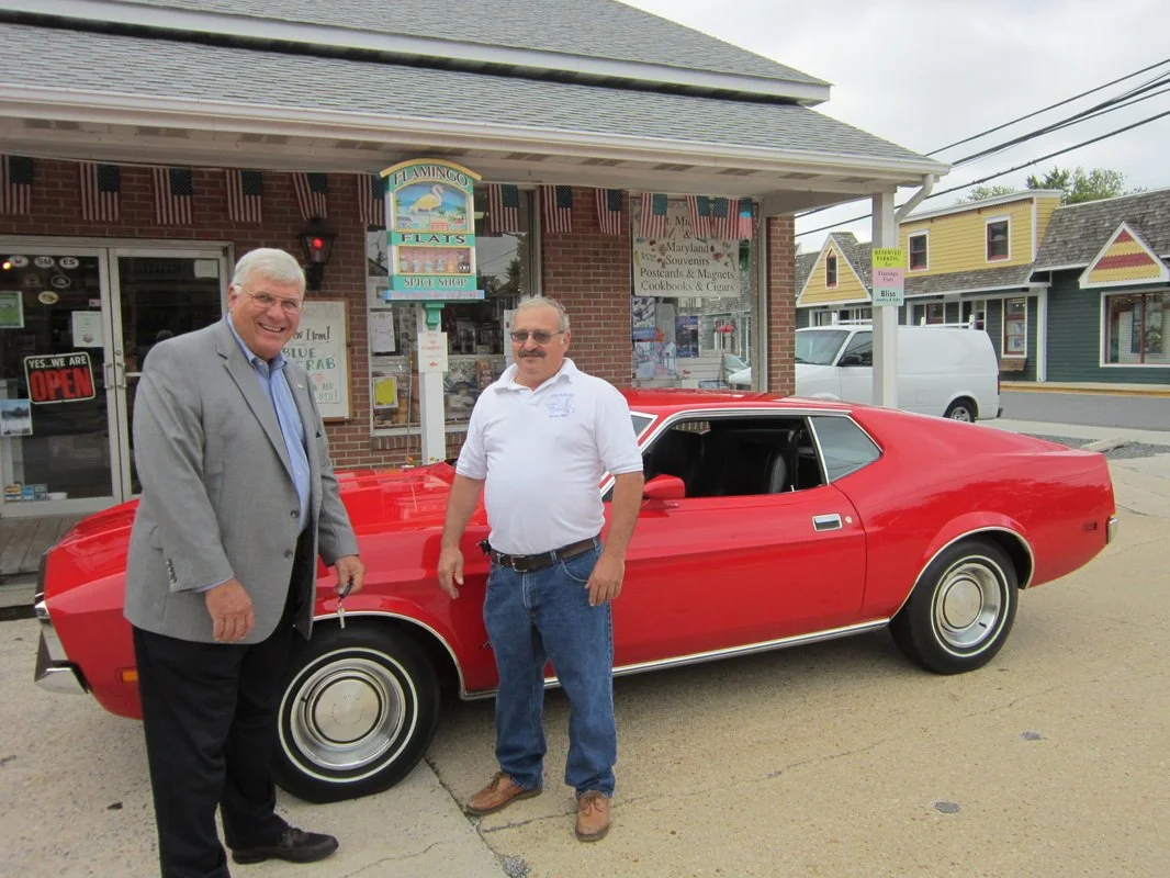 Two men standing in front of a red vintage car, outside a small shop with signs and flags, in a small town.