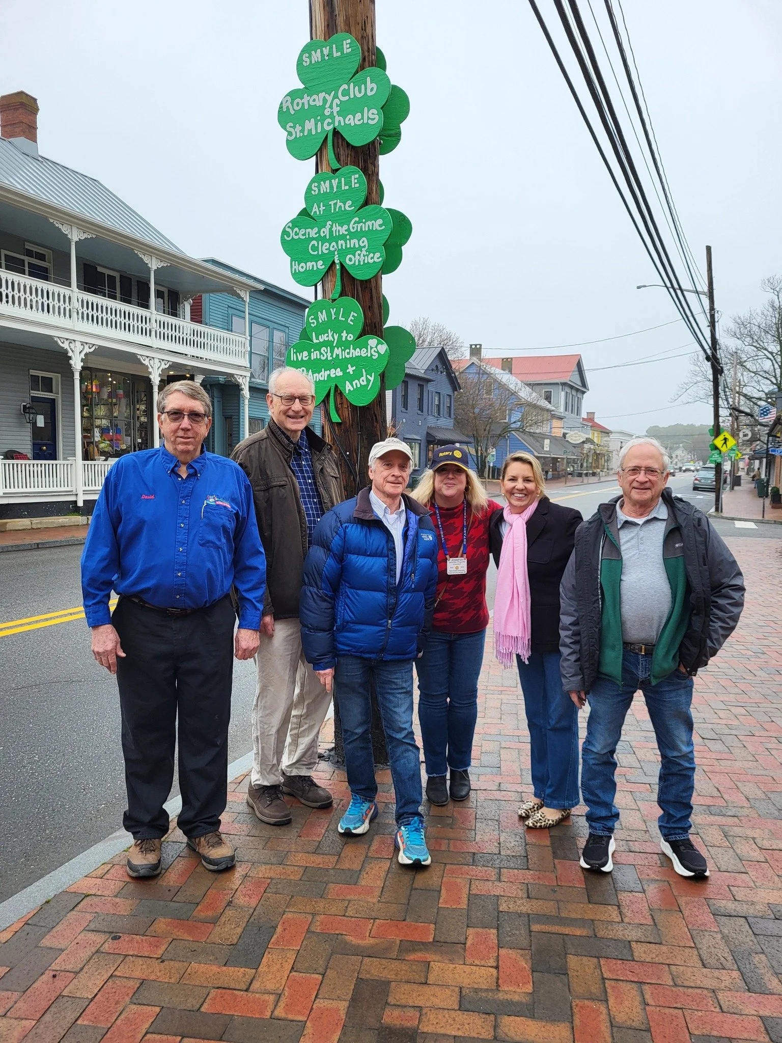 Six people standing on a sidewalk in front of a pole with green signs that read, 'SMYLE Rotary Club of St. Michaels,' 'SMYLE At The Scene of the Crime Cleaning Home & Office,' and 'SMYLE Lucky to live in St. Michaels.' The group is smiling and poses 