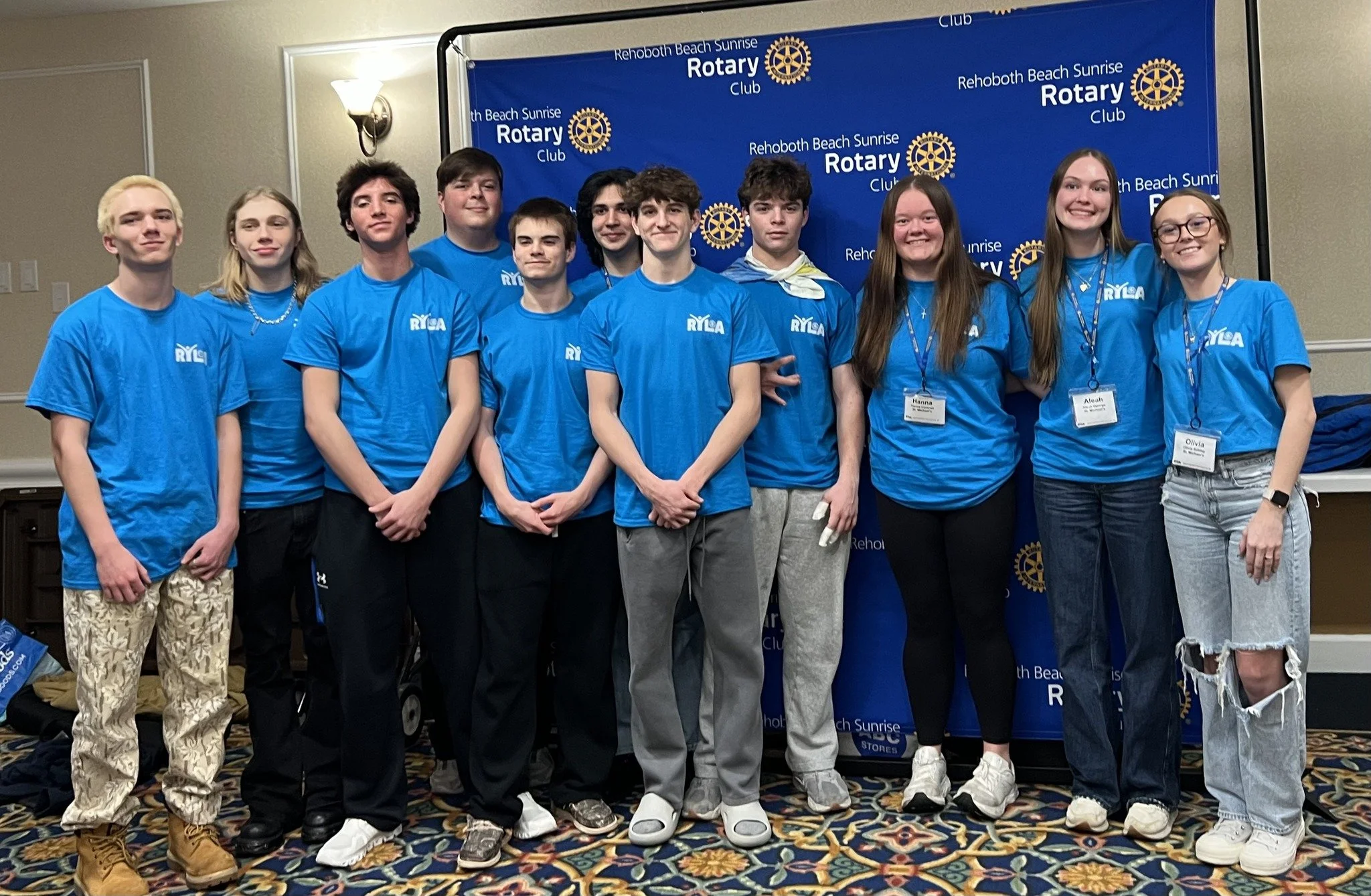 Group of young people wearing blue T-shirts with 'RYLA' logo, standing in front of a Rotary Club backdrop, posing for a photo at a conference or event.