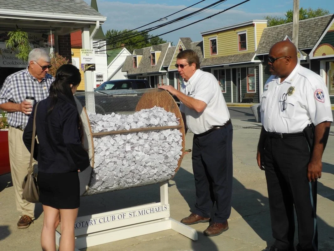 A group of five people gathered around a collection drum filled with paper tickets, outside on a sidewalk with colorful small houses in the background. Two men are in white shirts, one with a badge, and a woman in black with a bag, along with an olde