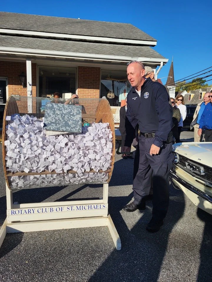 A man in a dark uniform stands next to a large wooden raffle drum filled with crumpled paper slips. The drum has a sign that reads 'Rotary Club of St. Michaels.' There are several people in the background, and a building with a porch behind them. The