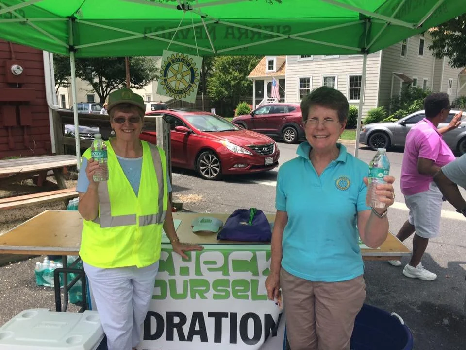 Two women smiling and holding water bottles under a green canopy at a community event, with a table and a Rotary Club sign.