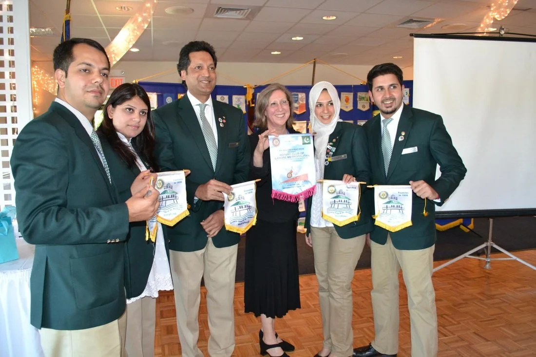 Group of six people, five men and one woman, dressed in green blazers and beige pants, at a formal event. The woman is holding a Rotary Club banner. They are standing in a room with a projector screen and a decorated background.